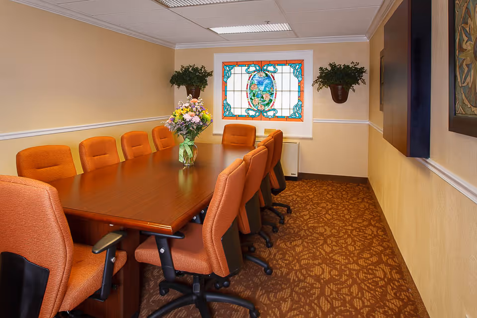 Conference/meeting room with a long wooden table, orange swivel chairs, a vase of flowers, and a stained-glass window.