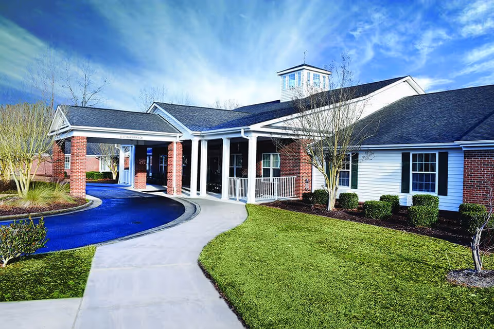 Exterior view of a senior living facility building with a covered entrance supported by brick columns, a curved driveway, well-maintained green lawn, and leafless trees under a partly cloudy blue sky.