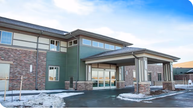 Exterior view of a two-story assisted living facility building with a covered entrance, brick and green siding, several windows, and a clear blue sky above. Snow is visible on the ground around the driveway.