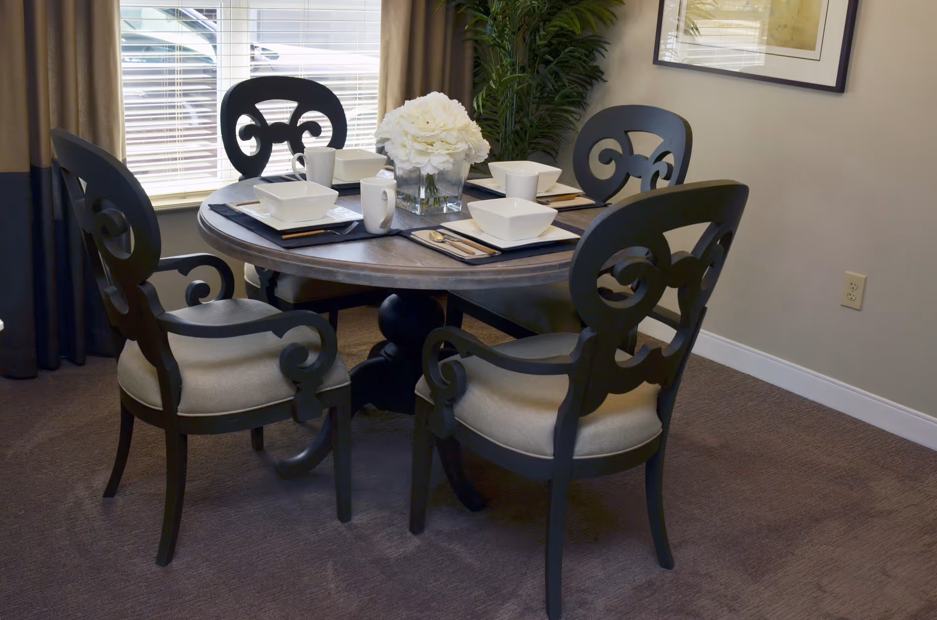A round dining table set for four with white square bowls, mugs, and silverware on black placemats. The table has a centerpiece of white flowers in a clear vase. Four black chairs with beige cushions surround the table. A window with blinds and a green plant are visible in the background.
