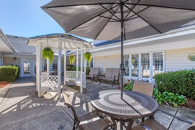 Outdoor patio area at Brookdale Utica Memory Care featuring a round metal table with an umbrella and chairs, a white gazebo with hanging plants, benches along the building wall, and well-maintained shrubs and flowers under a clear blue sky.