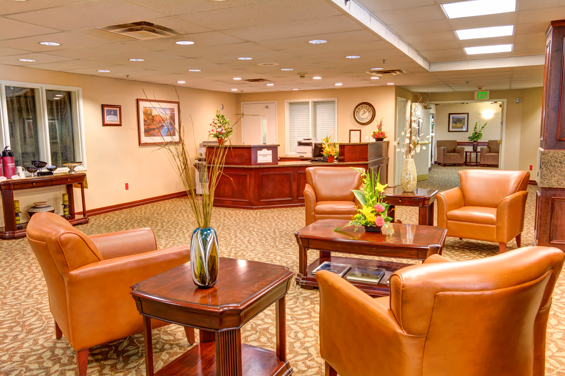 Reception lobby with leather armchairs, wooden tables, floral arrangements, and a front desk in the background.