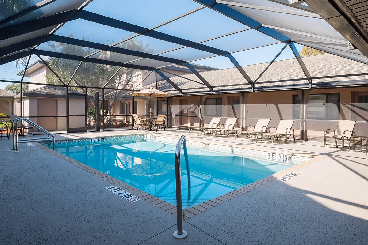 Indoor swimming pool area with a screened enclosure, surrounded by lounge chairs and a table with an umbrella. The pool has metal handrails and depth markers on the edge. The area is part of a residential or senior living facility with beige walls and windows in the background.