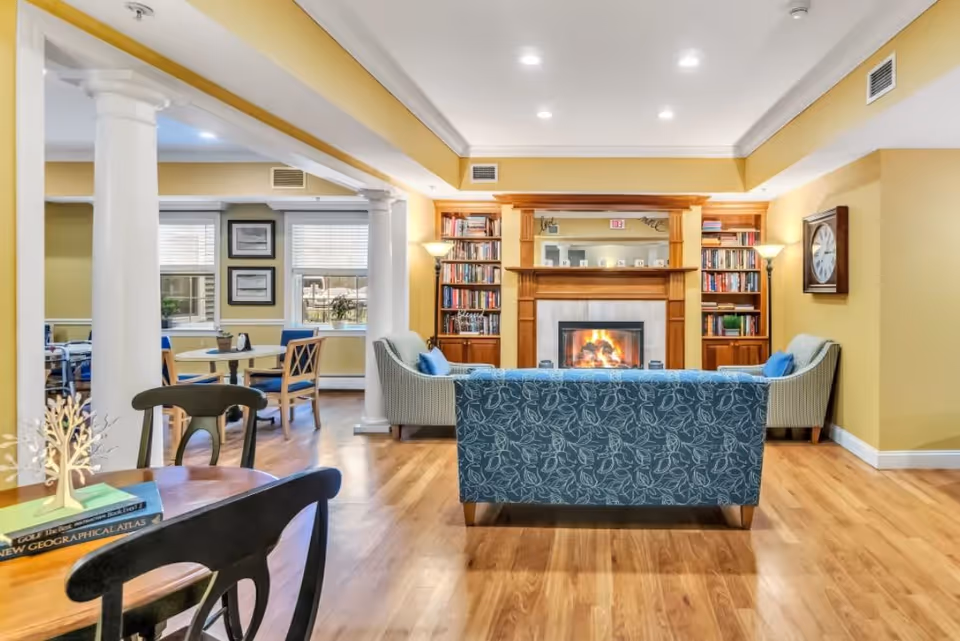 A cozy living room area with a blue patterned sofa and two armchairs facing a lit fireplace with a wooden mantel. The room features built-in bookshelves on either side of the fireplace, a large wall clock, and warm yellow walls. There are white columns separating the living area from a dining space with tables and chairs near windows with blinds. The floor is wooden, and the ceiling has recessed lighting.