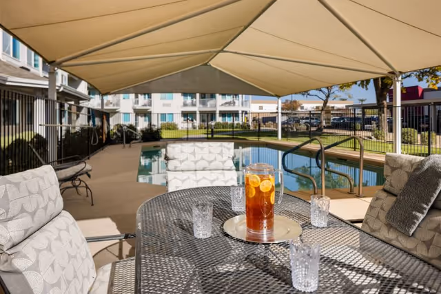 Outdoor seating area with cushioned chairs around a metal table holding a pitcher of iced tea with lemon slices and four empty glasses, situated next to a swimming pool under a large beige canopy. In the background, there is a fenced pool area, green lawn, trees, and a multi-story building.