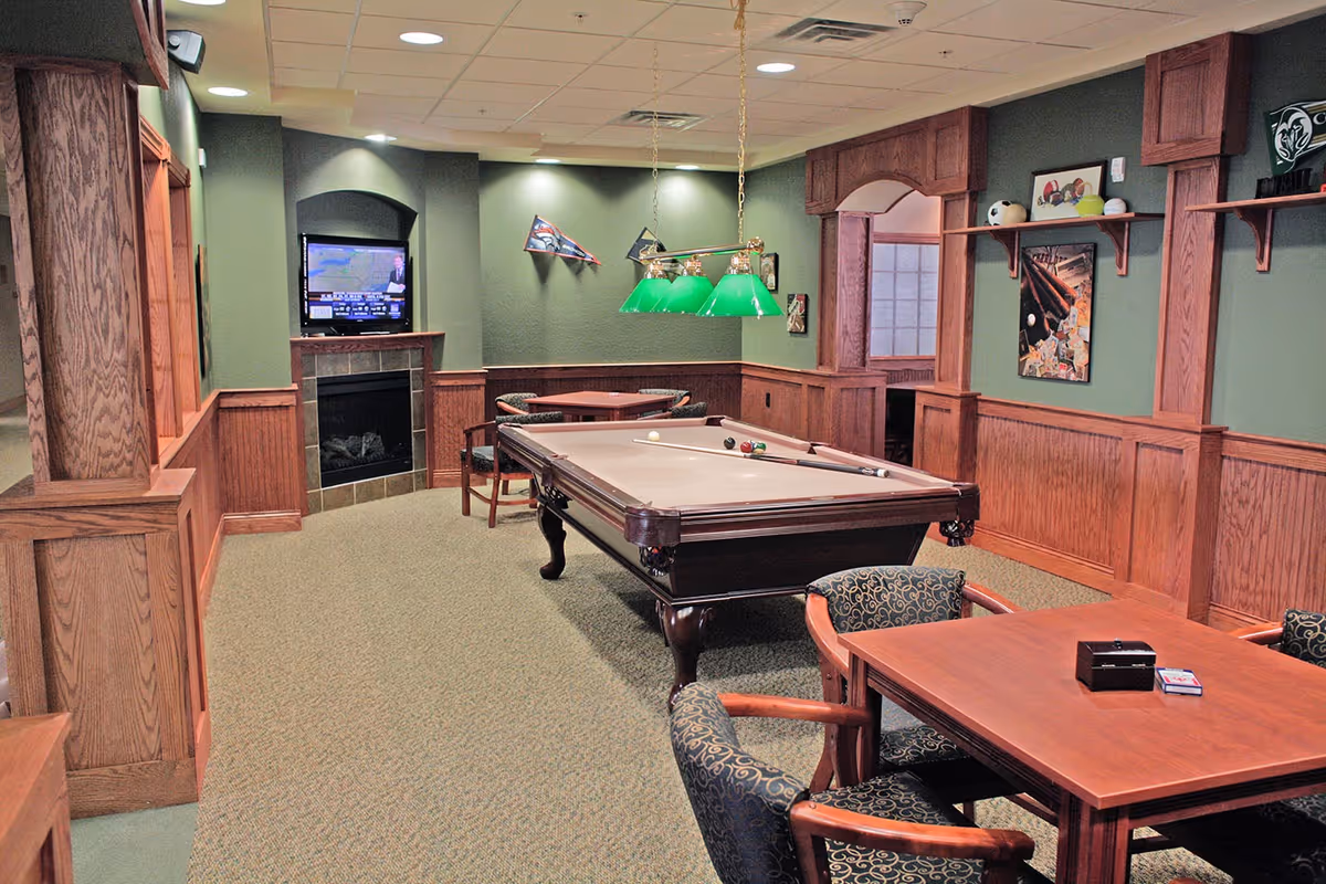 Interior recreation room with a pool table, seating area, TV above a fireplace, and wood-paneled green walls.