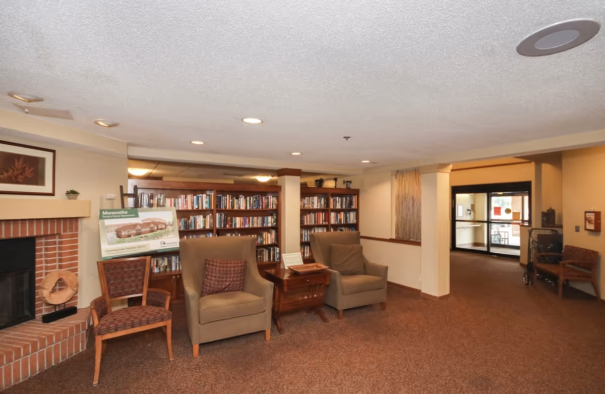 A cozy common area in Maranatha Place featuring two armchairs and a wooden chair arranged around a small wooden table. Behind the chairs is a bookshelf filled with books. To the left, there is a brick fireplace with a decorative wreath. The room has carpeted floors, recessed ceiling lights, and a doorway leading to another area with glass doors.