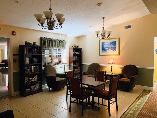 A cozy living room area in an assisted living facility with a square table and four chairs in the center. There are three armchairs arranged around two small side tables with lamps. Two tall bookshelves filled with books are against the walls, and a window with green curtains is visible. The walls are painted beige with white trim, and two chandeliers hang from the ceiling. A framed painting is on the wall above one of the armchairs.