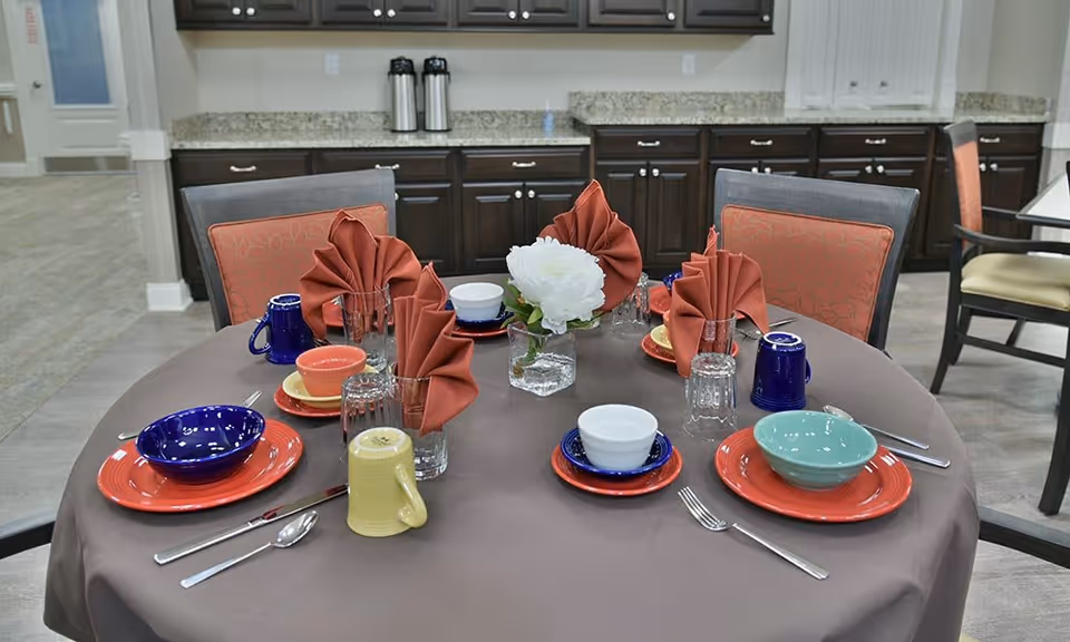 A round dining table set for six with colorful plates, bowls, cups, and folded rust-colored napkins. A small vase with a white flower is in the center. The table is covered with a brown tablecloth and surrounded by chairs with orange patterned cushions. In the background, there are dark wooden cabinets with a granite countertop.