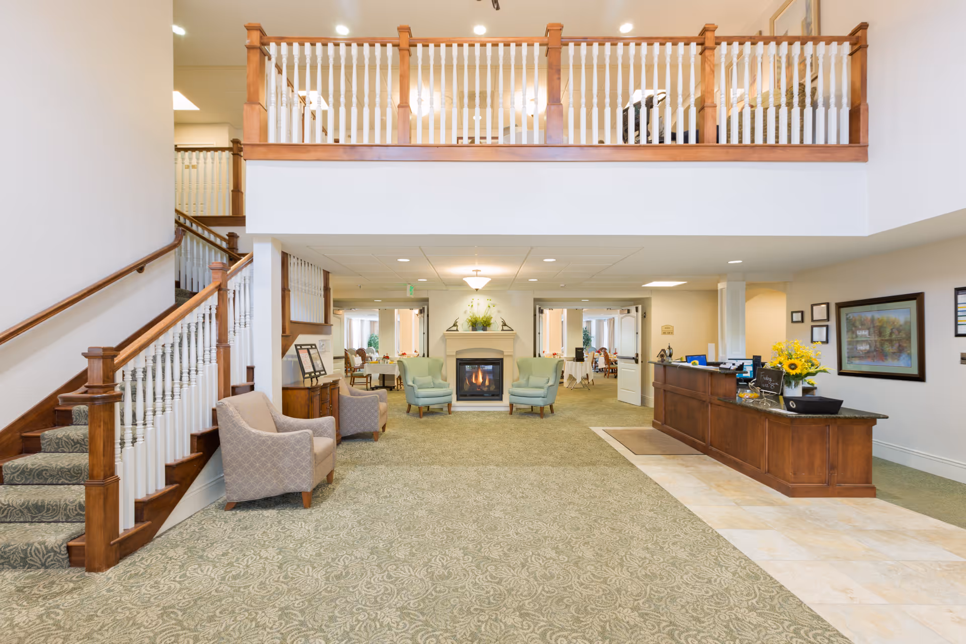 Spacious and well-lit assisted living facility lobby with a green patterned carpet, wooden staircase with white spindles, two armchairs near the stairs, a fireplace with two green armchairs in front, and a wooden reception desk with flowers and framed pictures on the wall.
