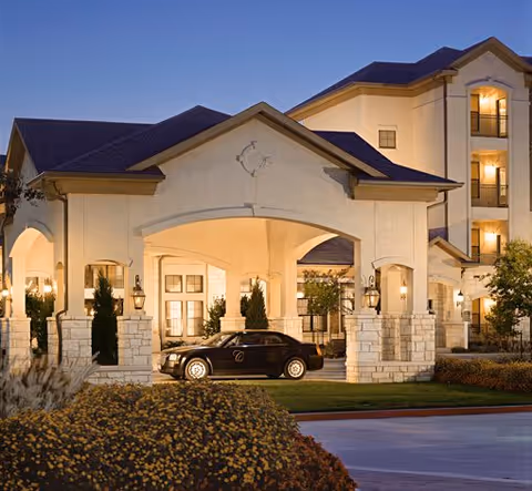 Exterior view of Conservatory At Keller Town Center building at dusk, featuring a covered entrance with stone pillars, warm lighting, and a black car parked under the entrance canopy.