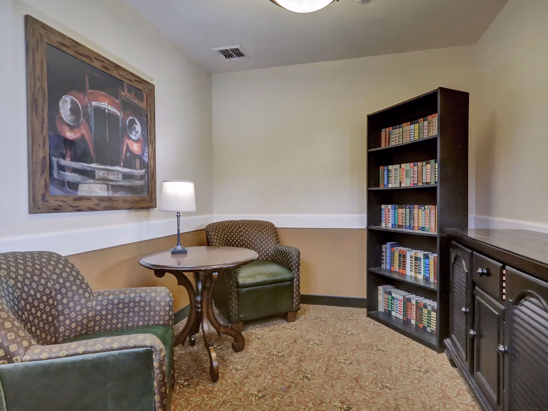 A cozy reading nook in a senior living facility featuring two patterned armchairs with green cushions, a round wooden table with a lamp on it, a tall bookshelf filled with books, and a framed painting of a vintage red car on the wall.