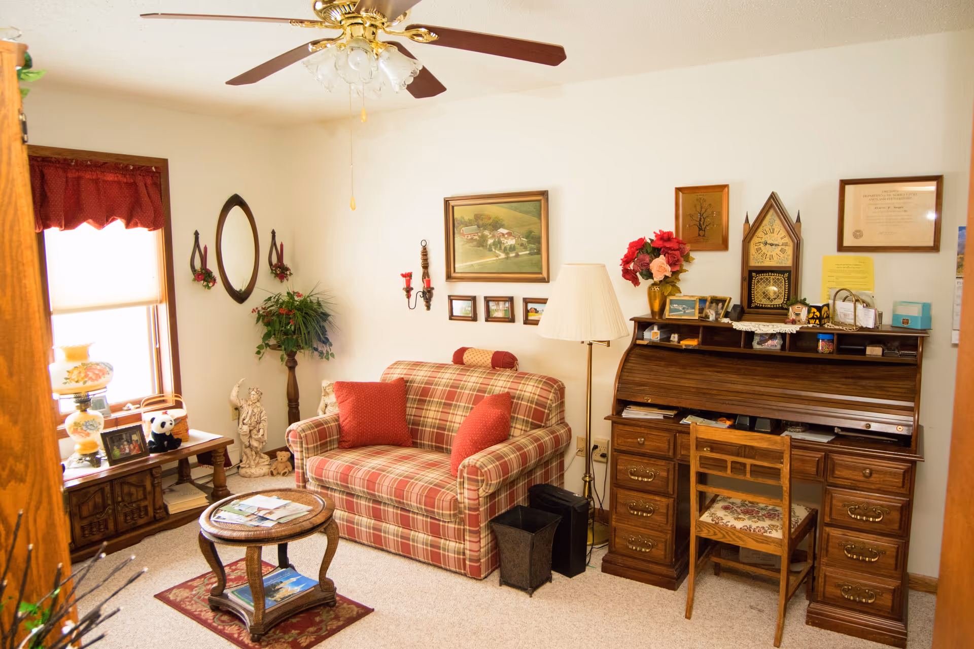 A cozy living room with a plaid loveseat adorned with red pillows, a wooden roll-top desk with a chair, a floor lamp, and various framed pictures and decorations on the walls. A small round wooden coffee table sits on a rug in front of the loveseat. There is a window with a red valance, a side table with a lamp and decorative items, and a ceiling fan with lights above.