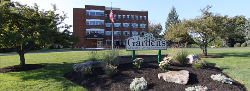 The front exterior of The Gardens Assisted Living Community building with a large sign in the foreground surrounded by landscaped plants and rocks, an American flag on a flagpole, and trees on a sunny day.