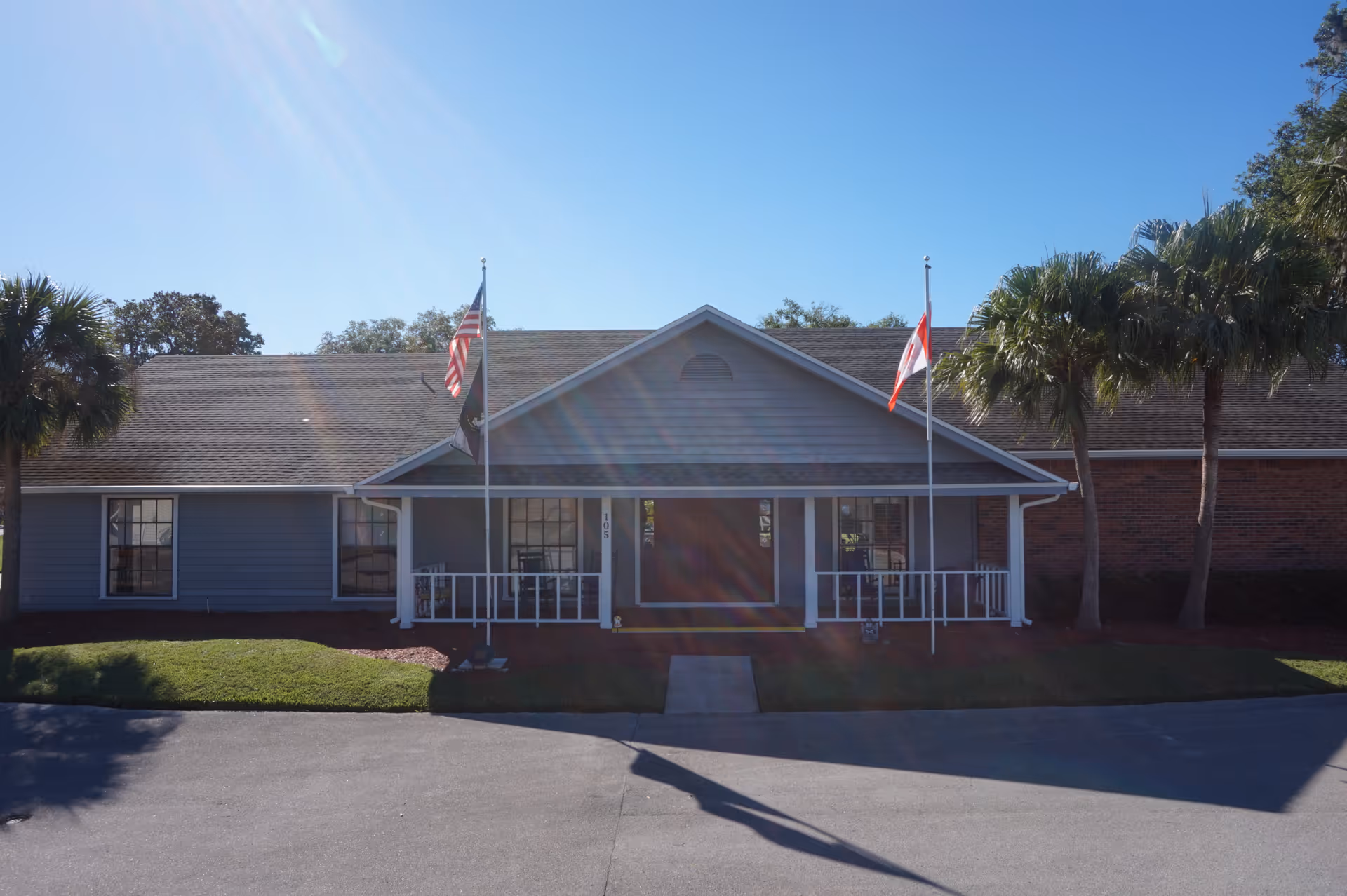 Front exterior view of a single-story building with a porch, two flagpoles displaying the American and Canadian flags, palm trees on either side, and a clear blue sky.