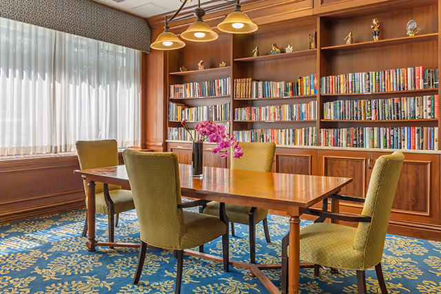 A cozy reading or meeting room with a wooden table surrounded by four green upholstered chairs. Behind the table is a large wooden bookshelf filled with books and decorative items. A vase with purple orchids is placed on the table. The room has a large window with sheer white curtains and a patterned blue and yellow carpet.