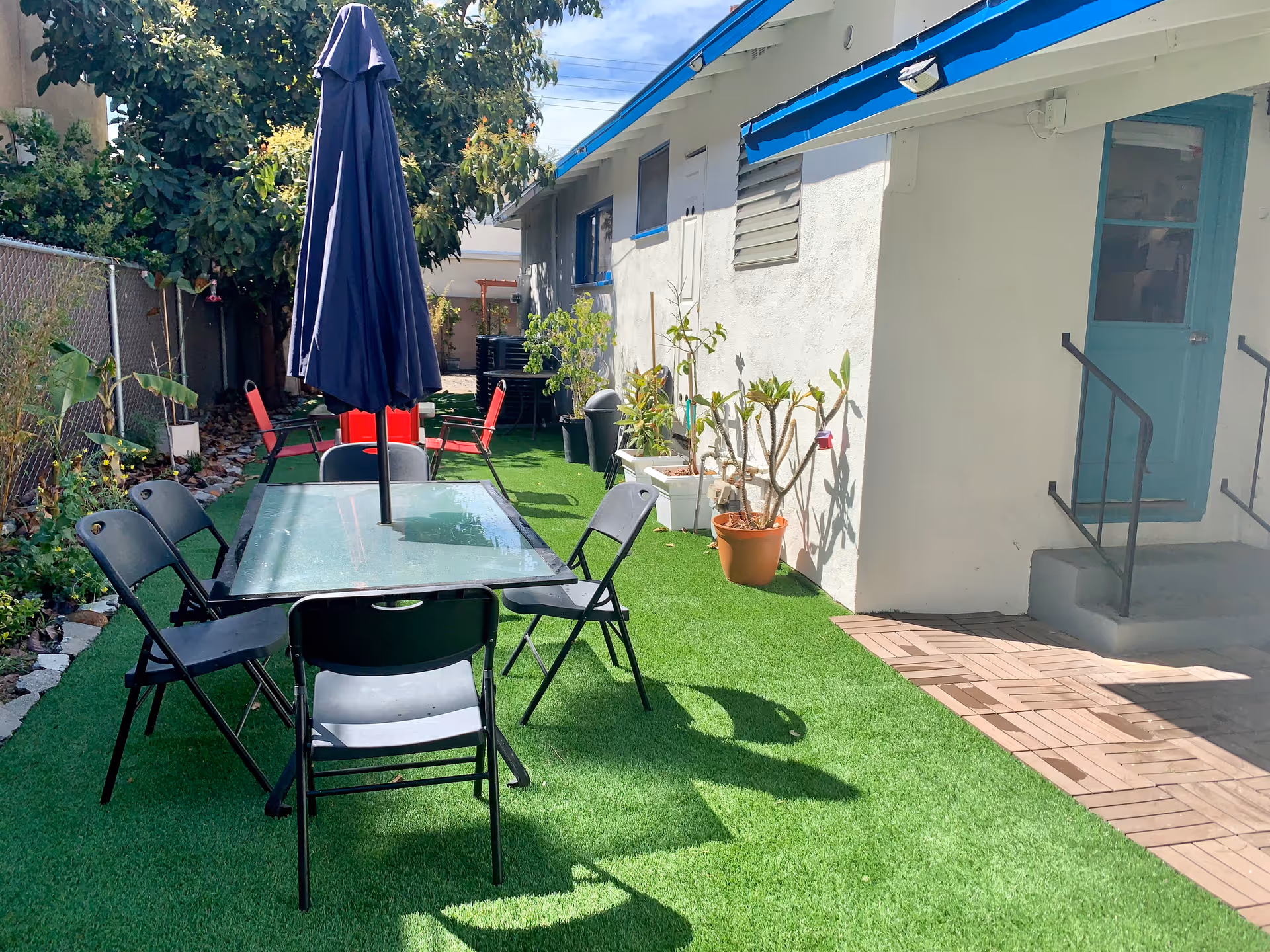 Outdoor patio area with artificial grass, a glass table with a closed navy blue umbrella in the center, surrounded by black folding chairs. There are potted plants along the side of a white building with a blue door and steps leading up to it. A chain-link fence and trees are visible on the left side.
