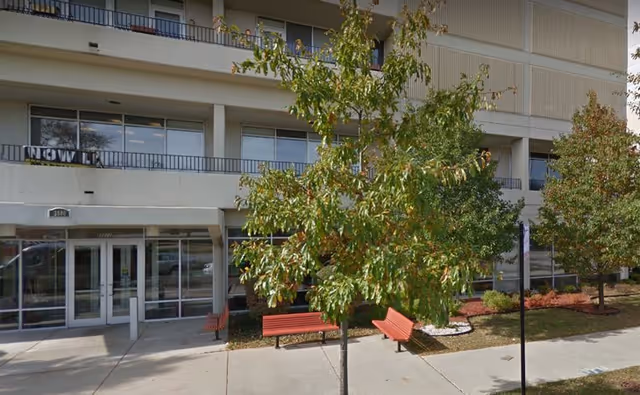 Exterior view of a multi-story building with balconies, trees, and red benches along a sidewalk in front of the entrance.