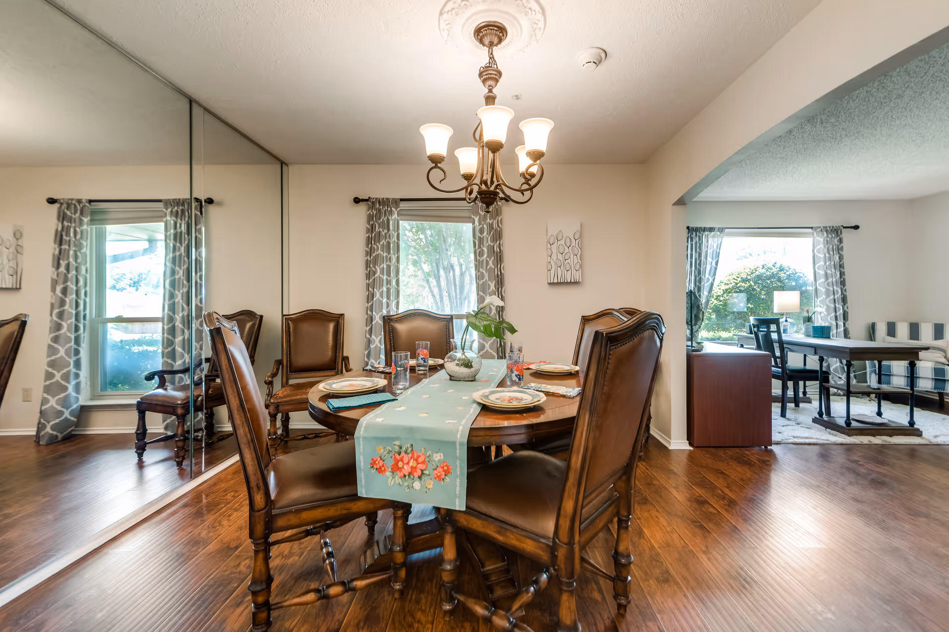A dining room with a round wooden table set for four with plates, glasses, and a floral table runner. The room has wooden flooring, a chandelier overhead, and large windows with patterned curtains. A large mirror covers one wall, reflecting the room. An adjacent living area with a window, chairs, and a lamp is visible through an open archway.