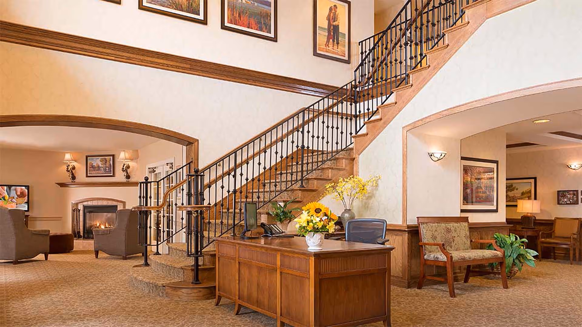 A warm and inviting senior living facility interior featuring a wooden reception desk with a computer and flower arrangements. Behind the desk is a staircase with a wrought iron railing leading to an upper floor. To the left, there is a cozy sitting area with armchairs and a fireplace, and to the right, additional seating with framed artwork on the walls.