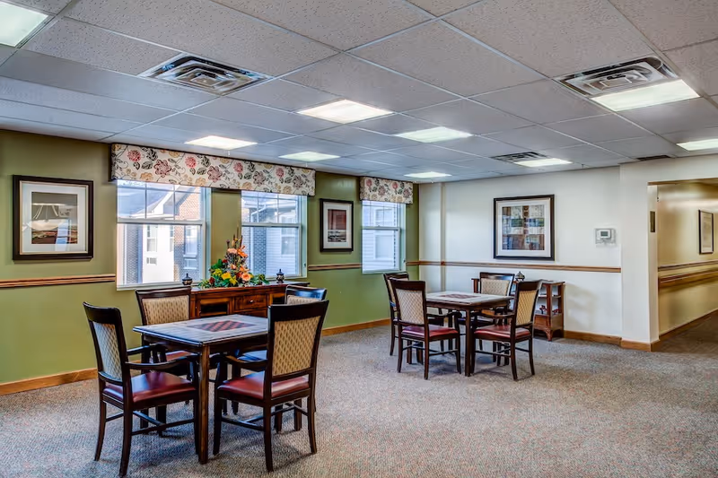 A well-lit dining area with two square wooden tables, each surrounded by four chairs with cushioned seats and backs. The room has green and white walls adorned with framed artwork and windows with floral valances. A wooden sideboard with a floral arrangement is positioned beneath the windows. The ceiling has recessed lighting and air vents.