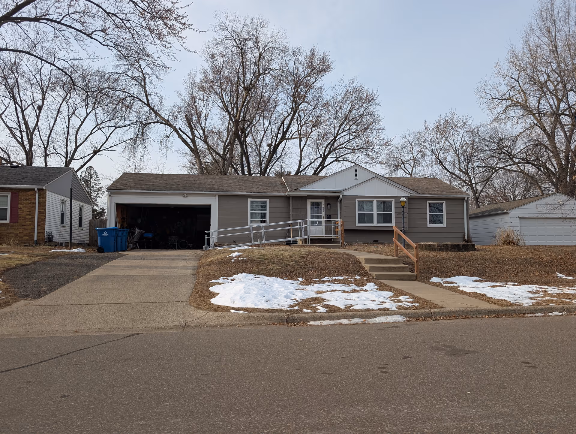 Front view of a single-story gray house with an open garage, a wheelchair ramp and front steps, bare trees, and patches of snow on the lawn.