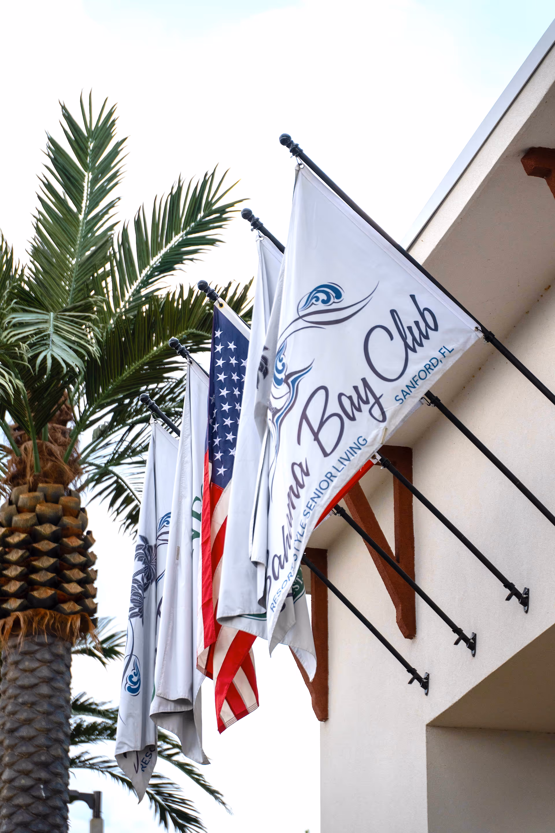 Flags mounted on poles attached to the exterior wall of a building, including an American flag and flags with the Bahama Bay Club logo, with palm trees visible in the background.