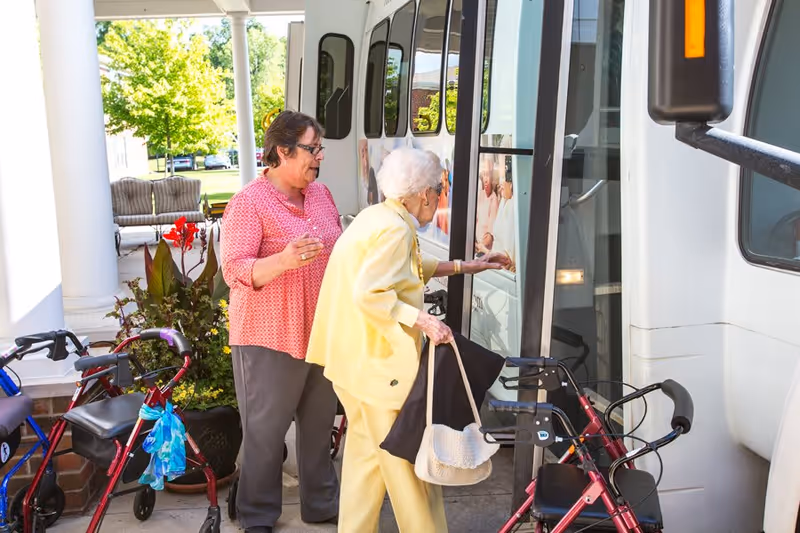 An elderly woman in a yellow outfit with a cane and handbag is boarding a white shuttle bus with assistance from a caregiver wearing a pink top. Several walkers are parked nearby, and there is greenery and outdoor seating in the background.