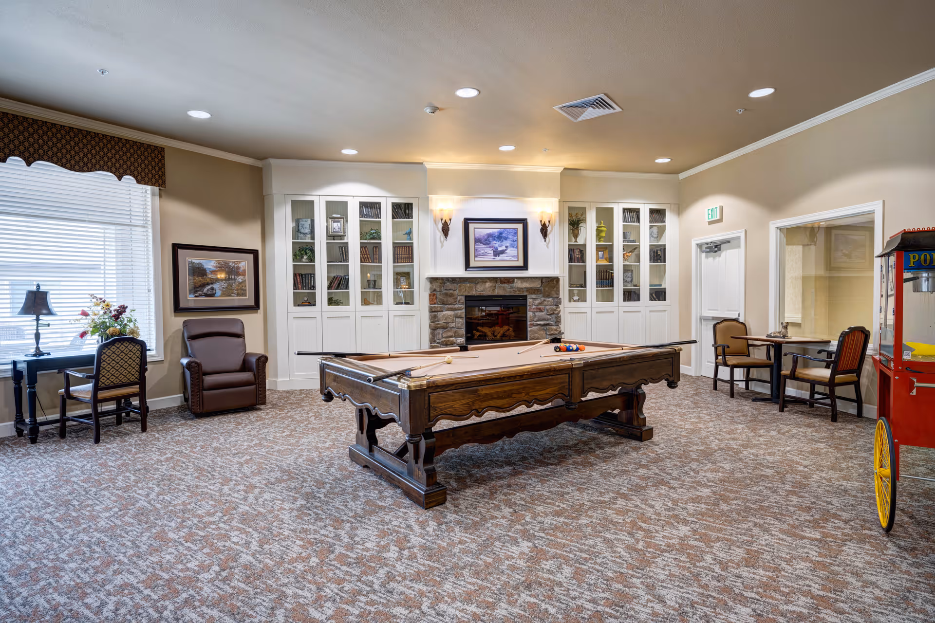 A spacious recreational room with a pool table in the center, a stone fireplace with built-in white cabinets filled with books and decorative items, a popcorn machine on the right, and seating areas including a leather armchair, a small table with chairs near a window, and another table with chairs near a door.