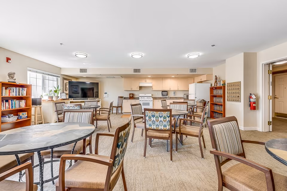 Communal dining and lounge area with round tables and chairs, a kitchenette and TV in a senior living facility.