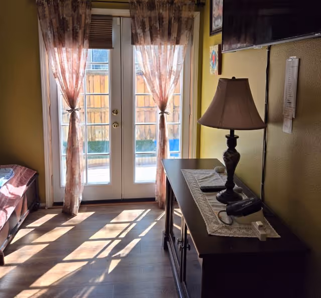 Sunlit bedroom with French doors, sheer pink curtains, and a dresser with lamp.