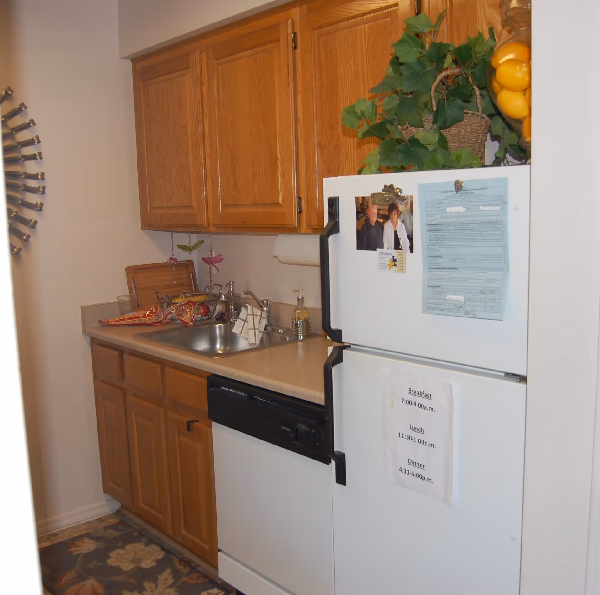 A small kitchen area with wooden cabinets, a countertop with a sink, and a white refrigerator. The refrigerator has papers and photos attached to it, and there is a plant and a bowl of yellow fruit on top. A dishwasher is located below the counter, and a decorative wall piece is partially visible on the left.