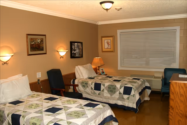 Shared bedroom with two twin beds covered in patterned quilts, chairs, a bedside lamp, and a window with blinds.