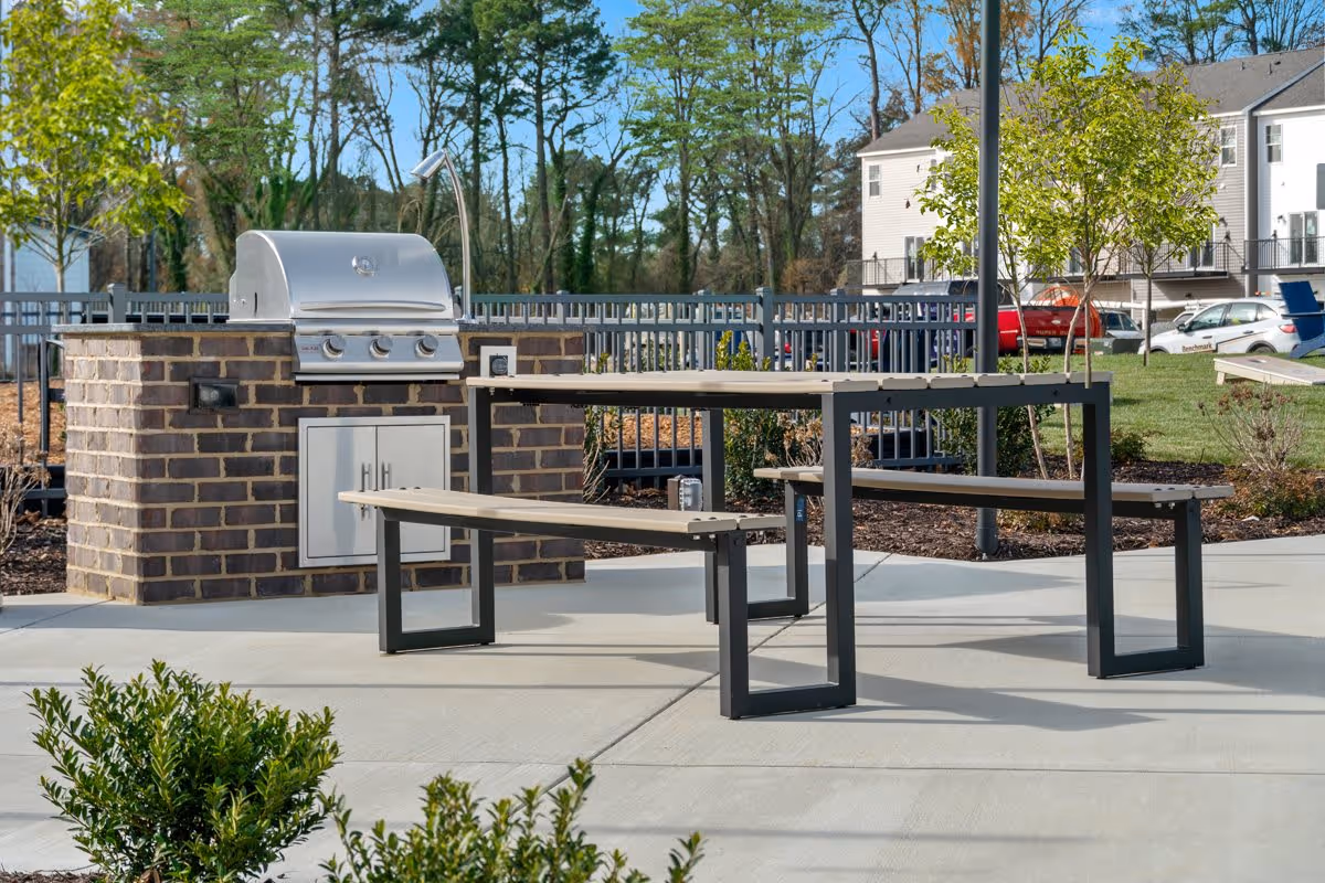 Outdoor patio with a built-in brick barbecue grill and picnic-style benches, with apartment buildings in the background.