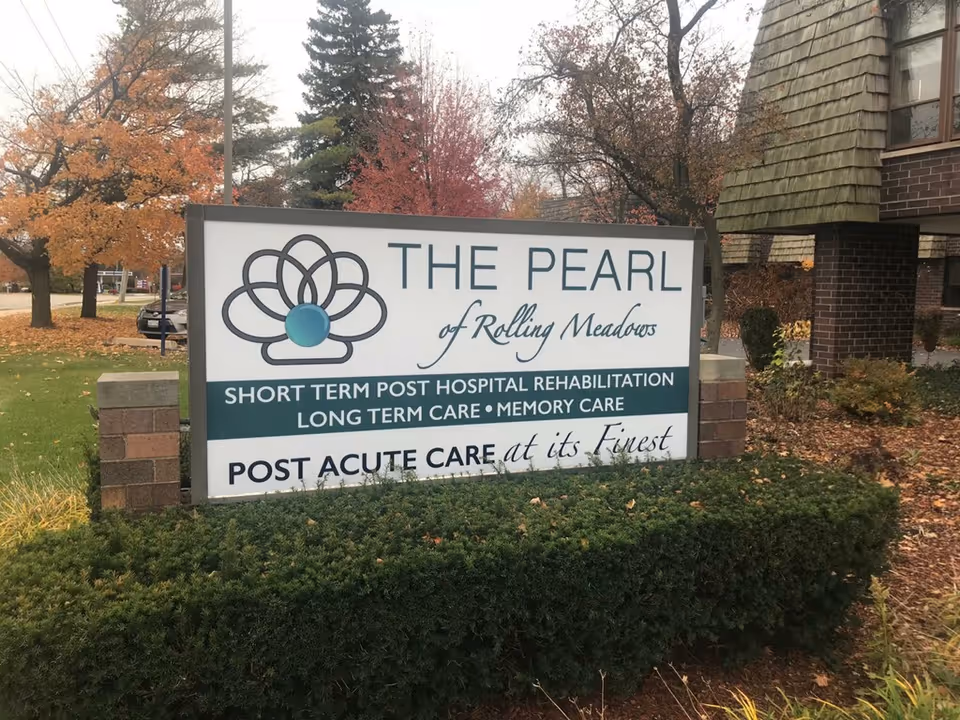 Outdoor sign for 'The Pearl of Rolling Meadows' on landscaped grounds in front of a brick building with autumn trees.