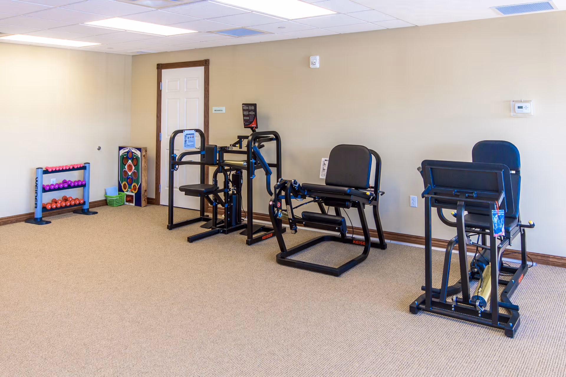 A small exercise room with beige walls and carpeted floor containing three pieces of black exercise equipment and a rack of colorful dumbbells in the corner near a closed white door.