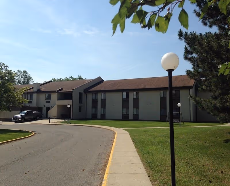 Exterior view of a two-story senior living facility building with a brown roof and beige walls. There is a curved driveway with a sidewalk and lamp posts along the path. Trees and green grass surround the building under a clear blue sky.