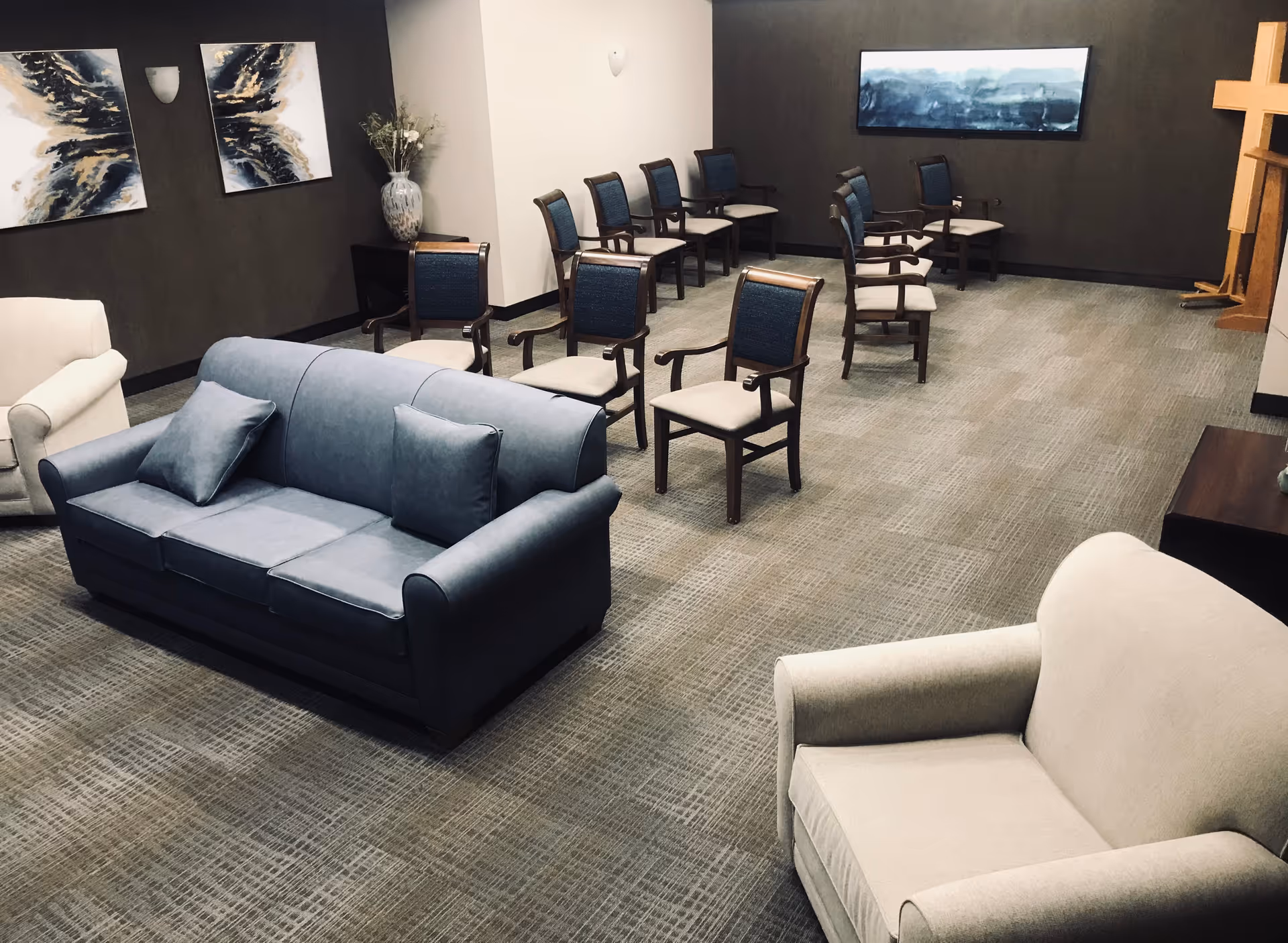 A spacious room with multiple chairs arranged in rows, two sofas, and a few armchairs. The walls are decorated with abstract paintings and a large cross is visible in the corner, suggesting a chapel or meeting room setting.