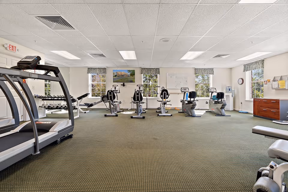 A well-lit fitness room with exercise equipment including treadmills, stationary bikes, and weight benches. The room has large windows with patterned valances, a green carpeted floor, and a white ceiling with fluorescent lights. There is a water cooler and a clock on the wall, along with some bulletin boards and a TV mounted on the far wall.