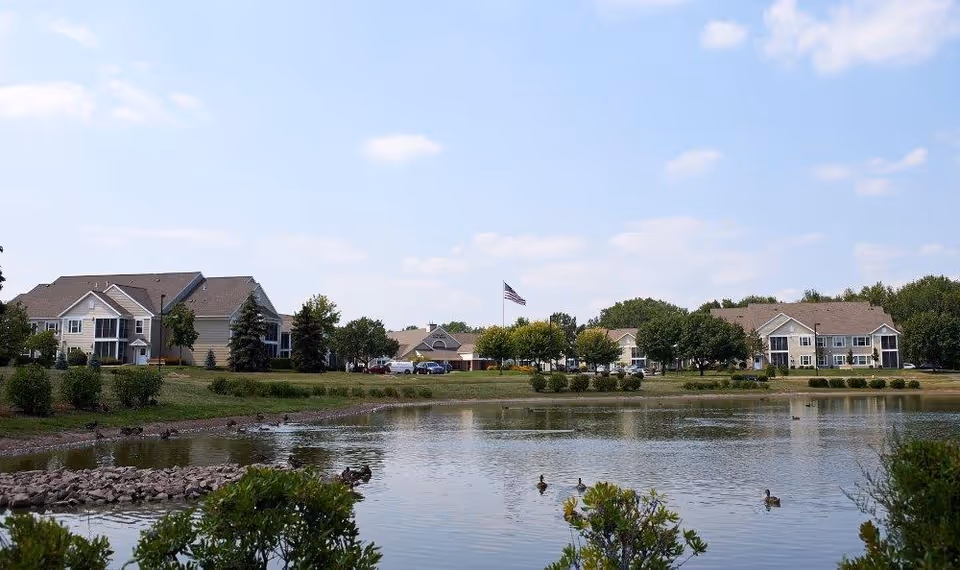 View of a senior living facility with multiple two-story buildings surrounding a pond with ducks swimming. There are trees, bushes, and a large American flag in the center of the scene under a partly cloudy sky.