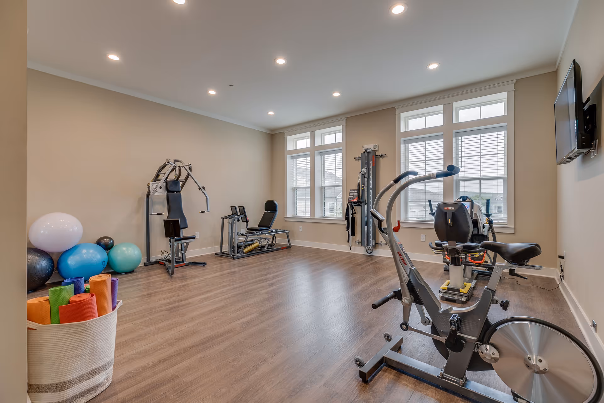 A well-lit exercise room with wooden flooring featuring various fitness equipment including a stationary bike, weight machine, exercise balls, yoga mats in a basket, and a wall-mounted TV. Large windows allow natural light to enter the room.
