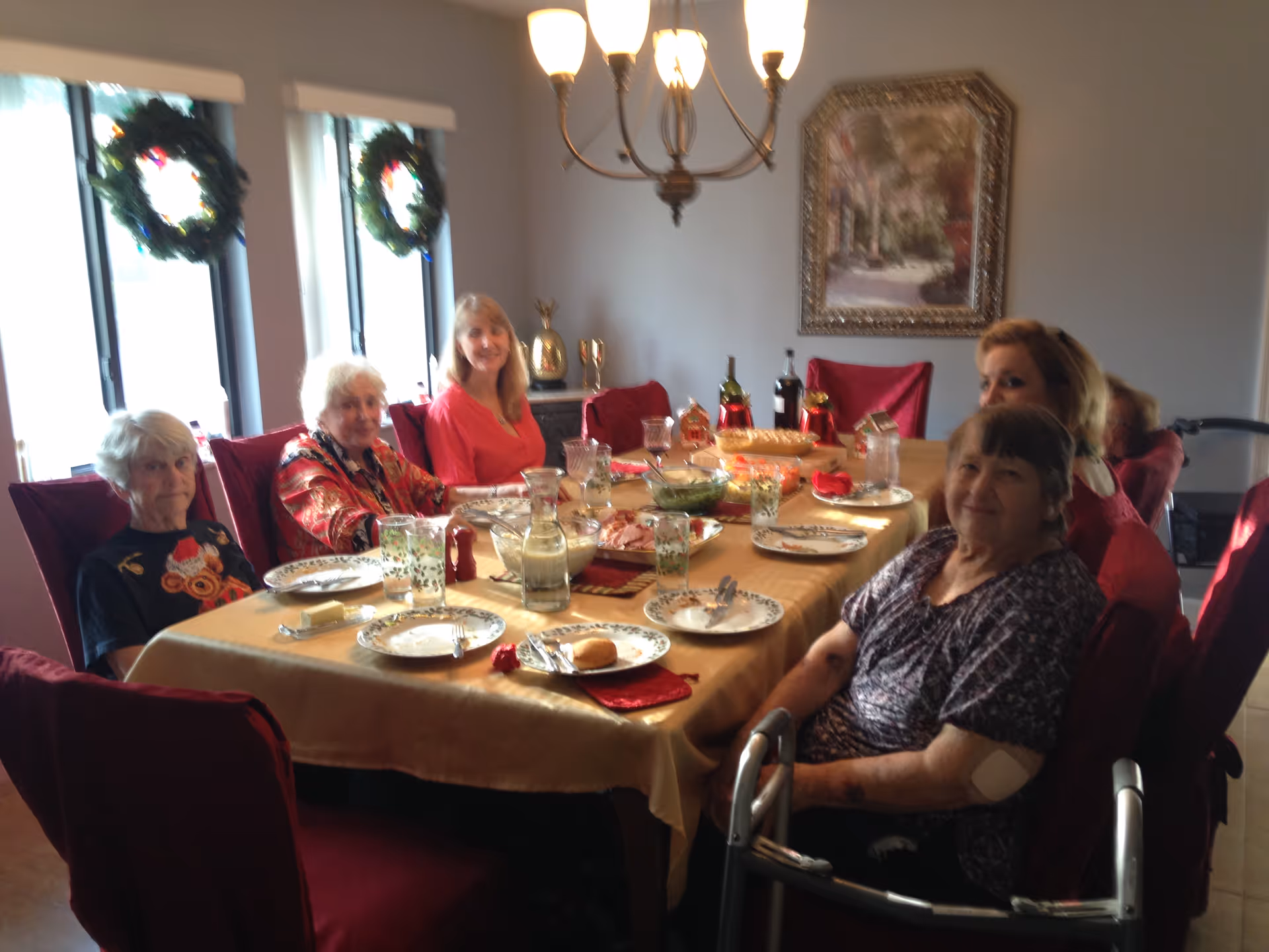 A group of six elderly women sitting around a dining table set with plates, glasses, and food in a room decorated with holiday wreaths on the windows and a chandelier overhead.