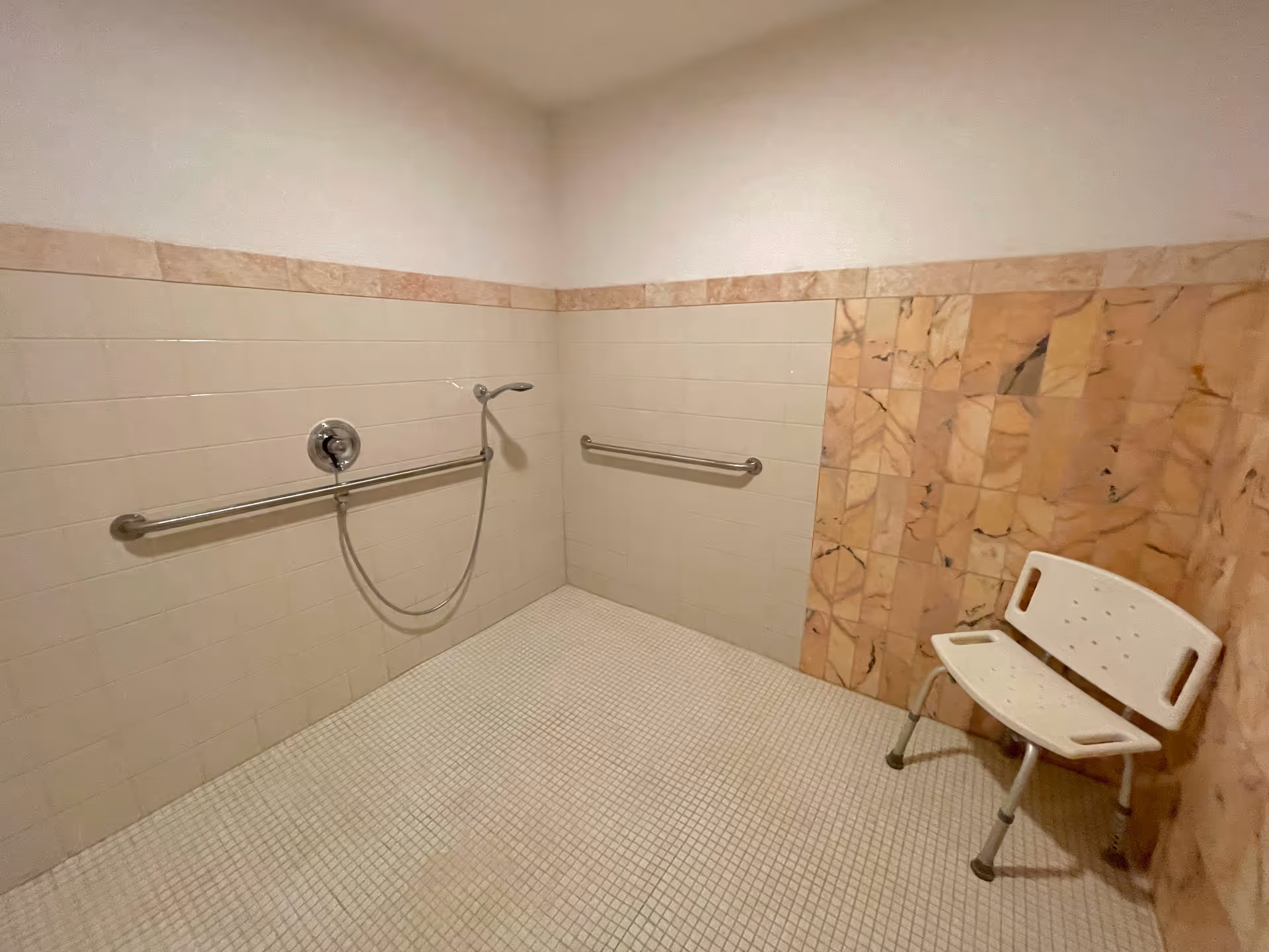 A tiled shower area with beige and light brown tiles, featuring two horizontal metal grab bars on the walls, a handheld showerhead attached to the wall, and a white plastic shower chair with metal legs positioned in the corner.