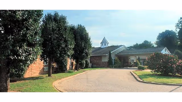 Exterior view of a single-story brick building with a small cupola on the roof, surrounded by trees and bushes, with a curved driveway leading to the entrance.