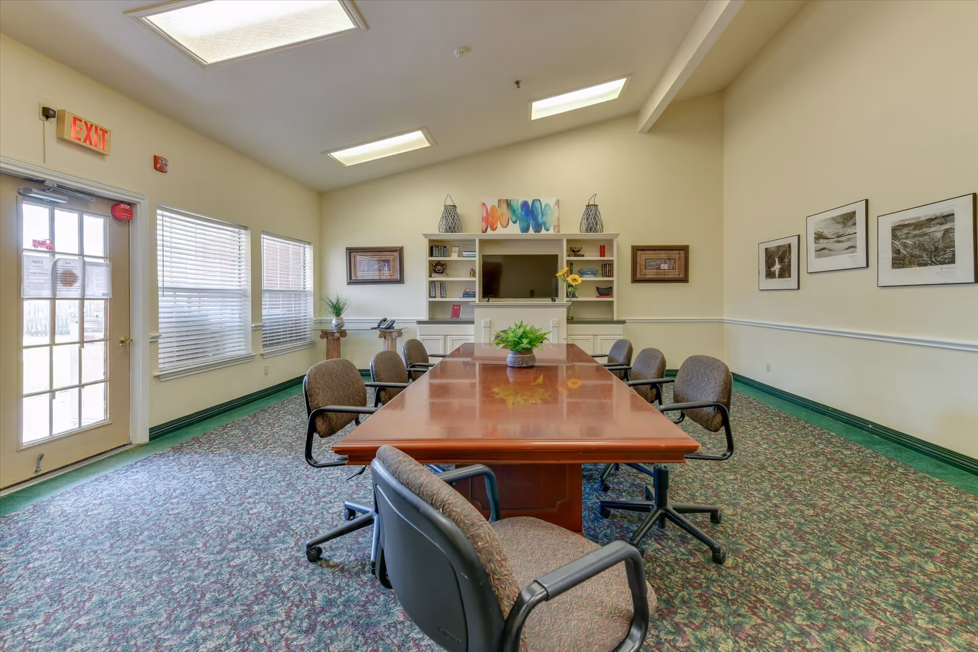 A conference or meeting room with a long wooden table surrounded by eight office chairs on a patterned carpet. The room has high ceilings with skylights, a wall-mounted TV, built-in shelves with decorative items, framed pictures on the walls, and windows letting in natural light. An exit door is visible on the left side.
