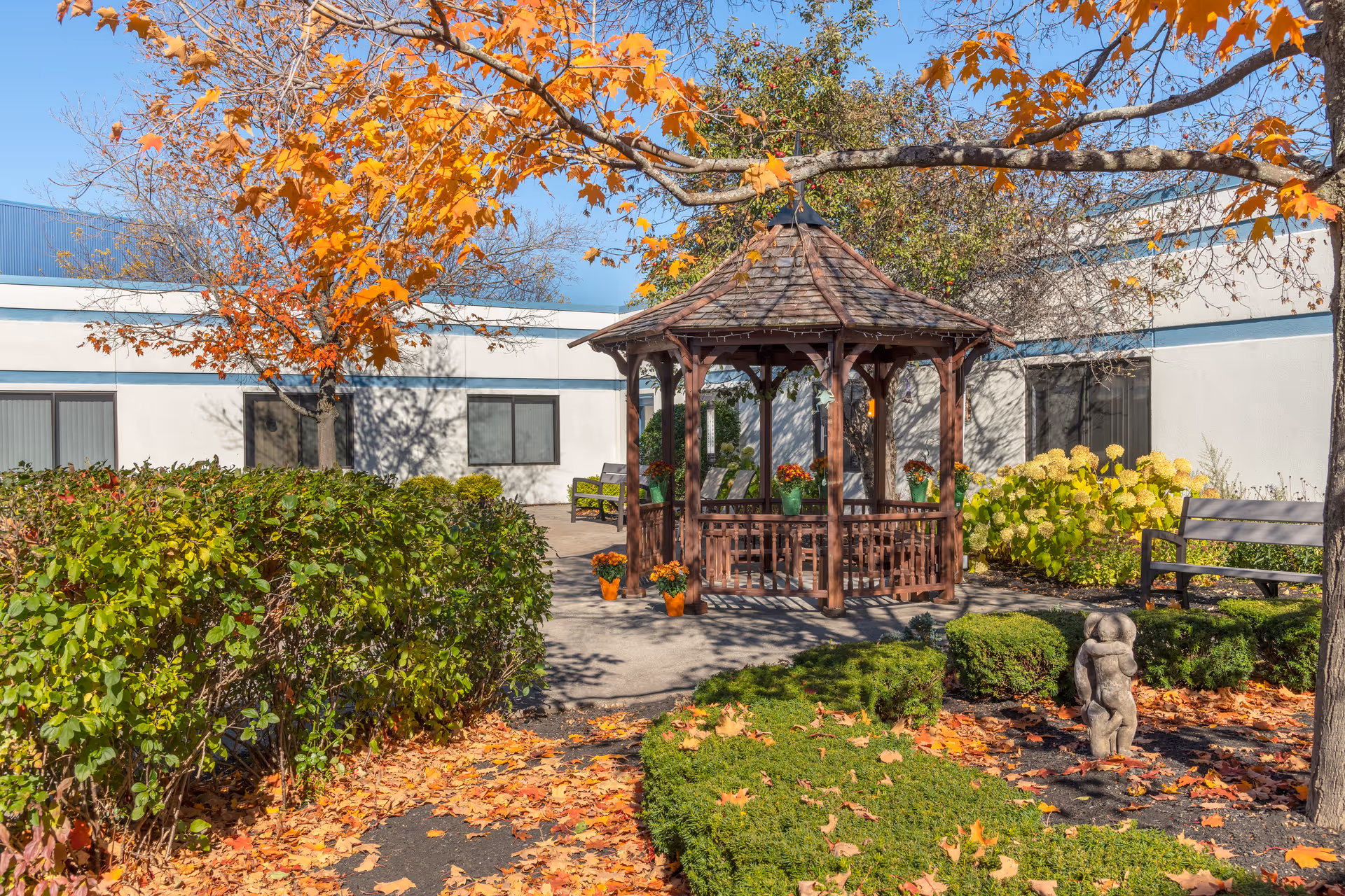 Outdoor courtyard featuring a wooden gazebo, benches, fall foliage, and landscaped shrubs in front of the facility.