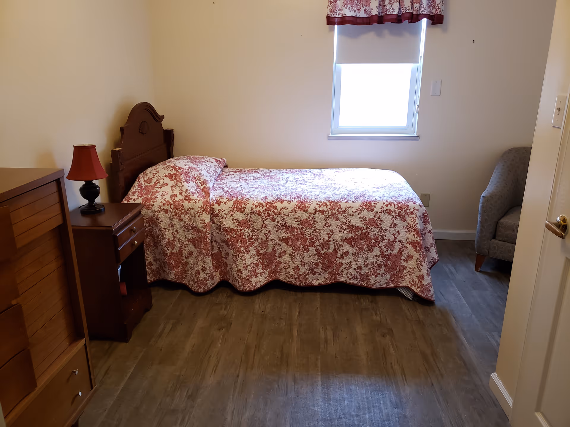 A small bedroom with a single bed covered in a red and white floral bedspread. There is a wooden headboard, a nightstand with a red lamp, a wooden dresser, a window with a floral valance, and a gray upholstered chair in the corner.