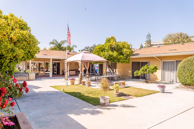Sunny outdoor courtyard with a gazebo, American flag, patio seating, and potted plants in front of single-story buildings.