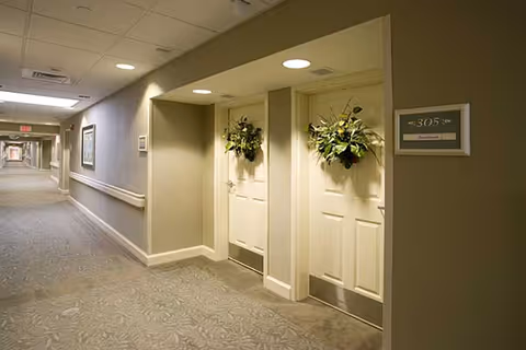 A well-lit hallway in a nursing center with beige walls and carpeted floor. Two white doors decorated with green leafy wreaths are visible on the right side, with a room number sign labeled 305 next to them. The hallway extends into the distance with handrails along the walls and framed pictures hanging.