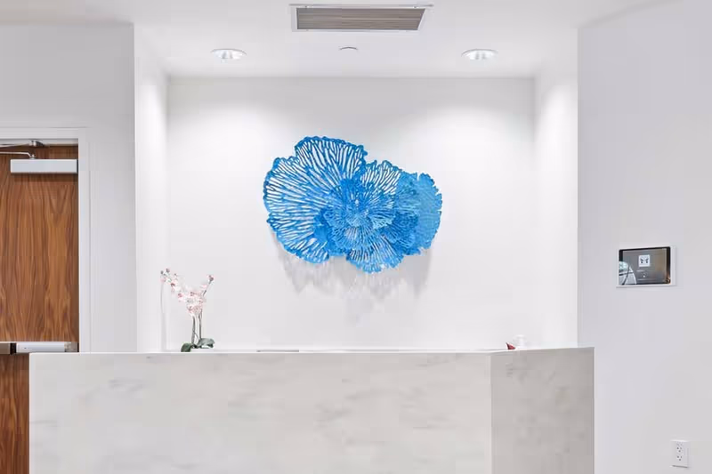 A modern reception desk with a white marble finish in front of a white wall. Above the desk is a blue decorative wall art piece resembling coral or abstract flowers. To the left is a wooden door, and to the right is a small digital screen mounted on the wall. A small vase with pink flowers is placed on the reception desk.
